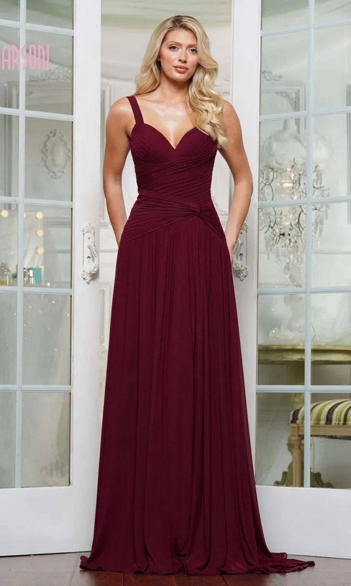 A woman poses indoors in front of glass doors, wearing the Marsoni MV1316 Long Formal Dress—a floor-length, sleeveless burgundy gown with a V-neckline and pleated detailing.